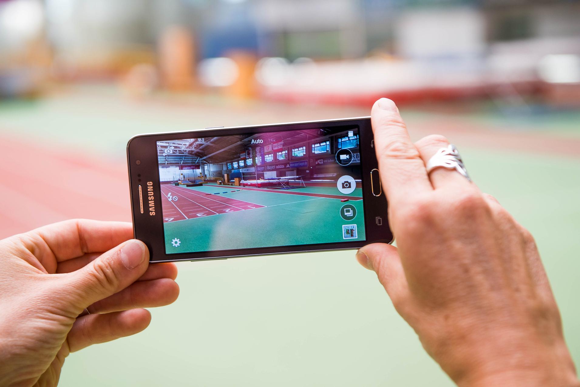 Hand hält ein Smartphone, das eine Sporthalle mit einer Laufbahn im Hintergrund zeigt.